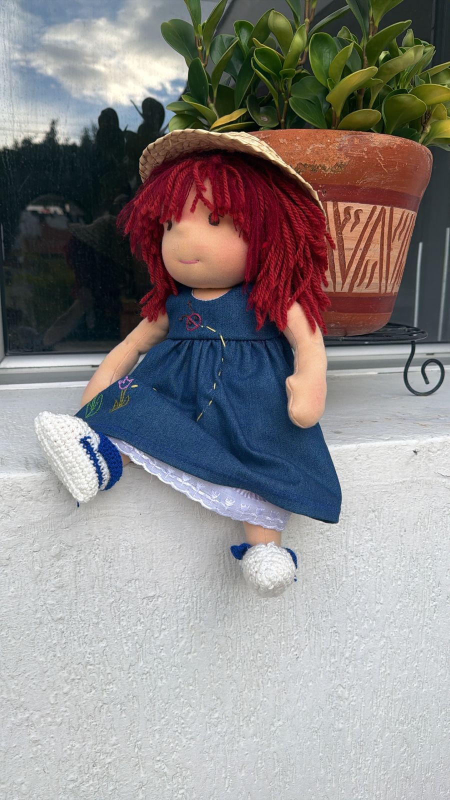 La misma muñeca con sombrero de paja, sentada junto a una matera.